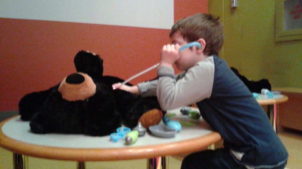 A small child pretends to do a medical exam on a teddy bear, using a plastic stethoscope. 